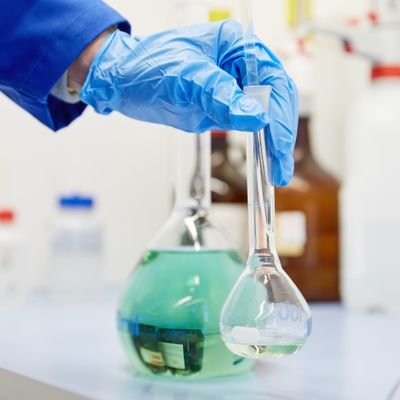 Blue-gloved hand holds a volumetric flask with clear liquid in a laboratory, with a larger flask of green solution in the background.